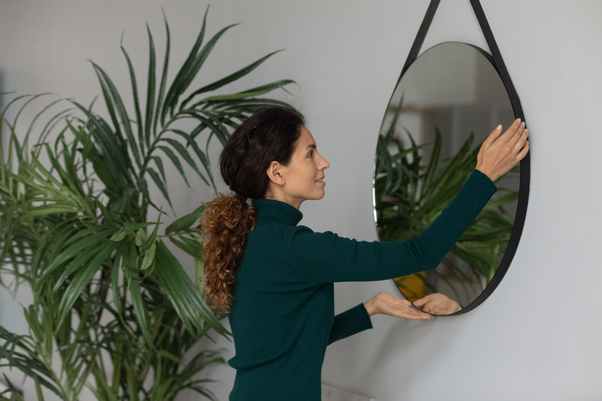 A woman with curly hair, wearing a dark green sweater, adjusts a round mirror on a white wall. There are green plants with large leaves to her left, adding a touch of nature to the indoor setting.