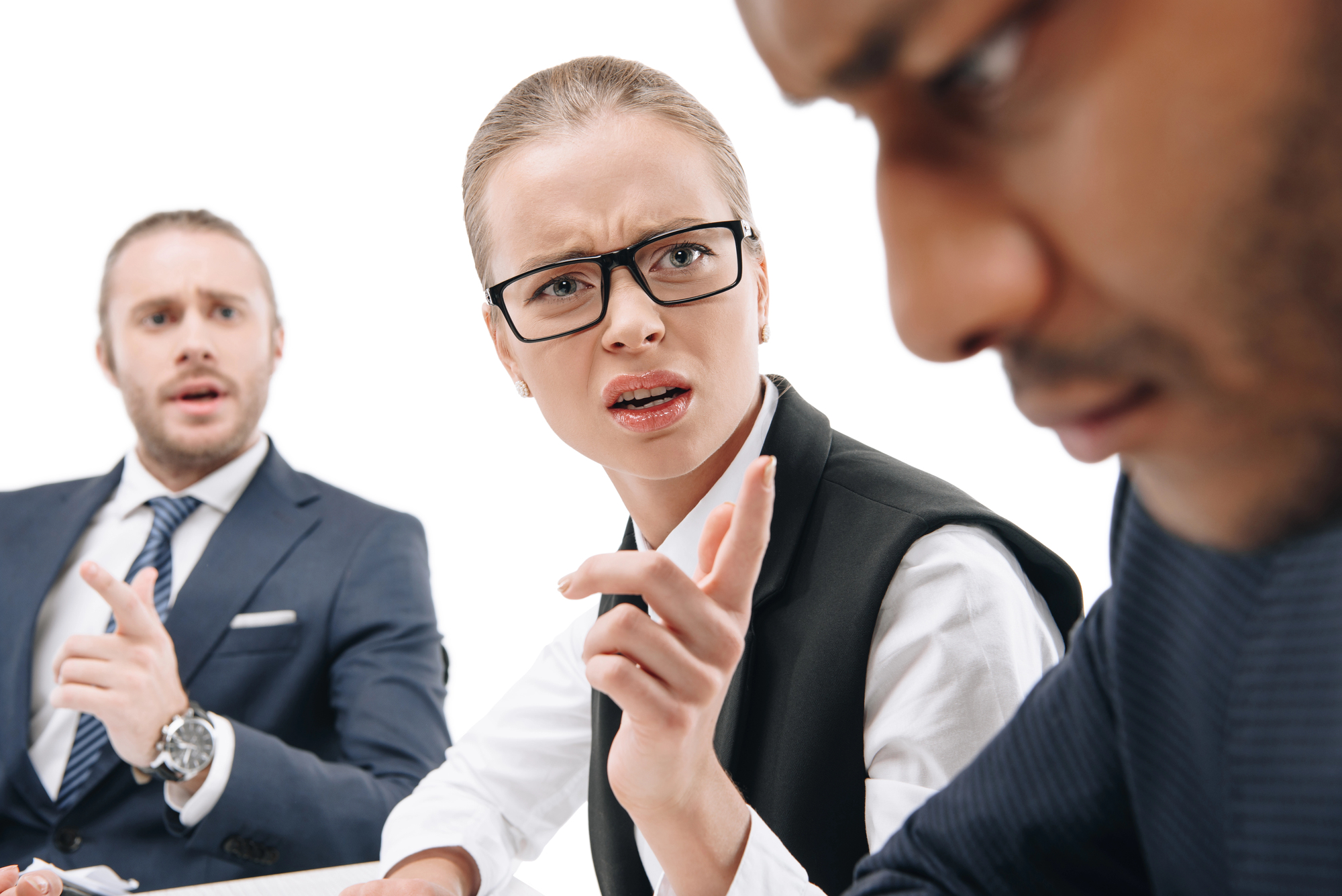 A woman in business attire, wearing glasses, looks directly at a man with an intense expression, while pointing her finger. Another man, also in business attire, sits beside her, appearing surprised. The background is white, creating a clean, professional setting.