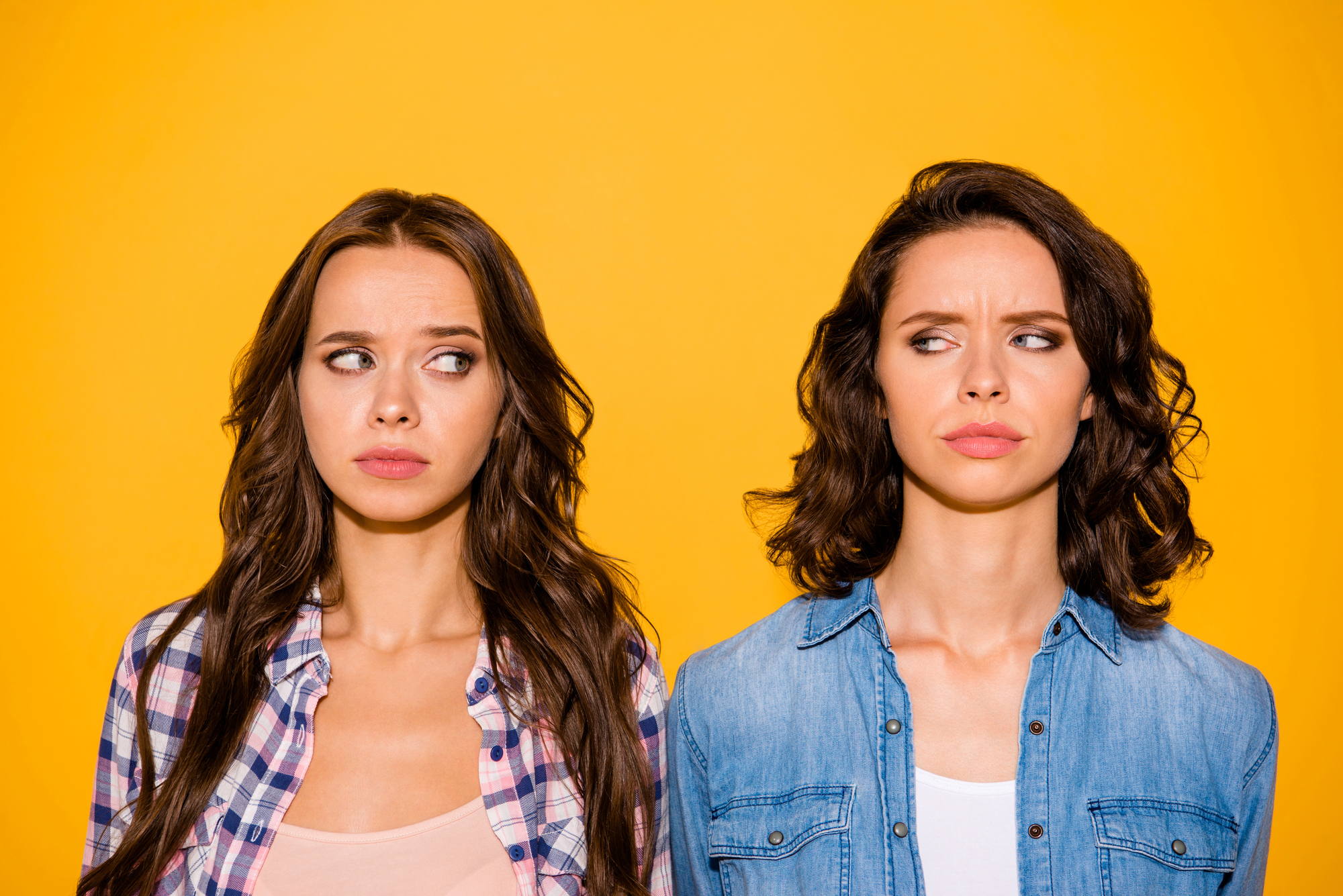 Two women stand side by side against a yellow background, both with skeptical expressions. The woman on the left has long, wavy hair and wears a plaid shirt, while the woman on the right has short, wavy hair and wears a denim shirt.