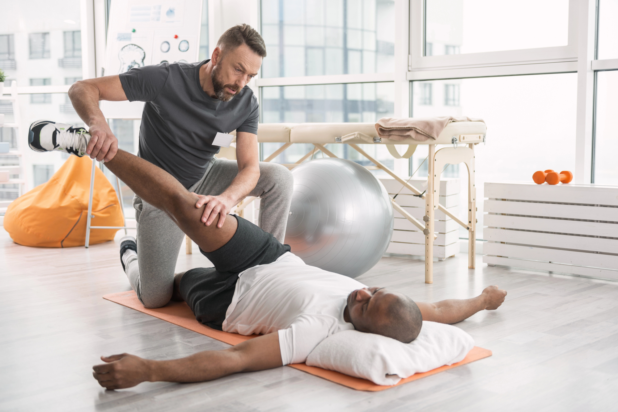 A physiotherapist assists a man lying on a mat with a leg stretch in a bright room. The therapist holds the man's leg while the man rests his head on a pillow. Exercise equipment, including a stability ball and dumbbells, are in the background.