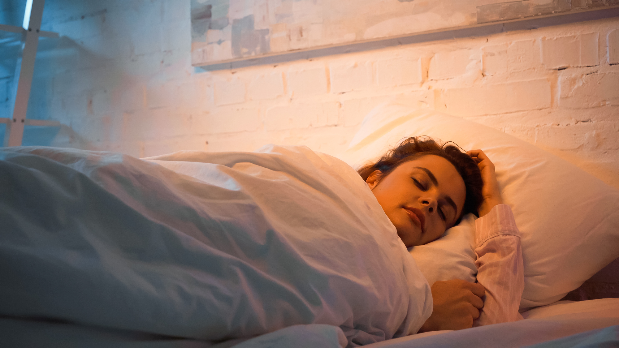 A person sleeping peacefully in a bed, covered with a white blanket, lying on a pillow. The room has a warm ambient light with a white brick wall and a soft, serene atmosphere.