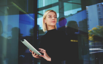 A woman in a black outfit stands confidently in front of a modern glass building, holding a tablet. The reflection of the cityscape and trees is visible in the background.