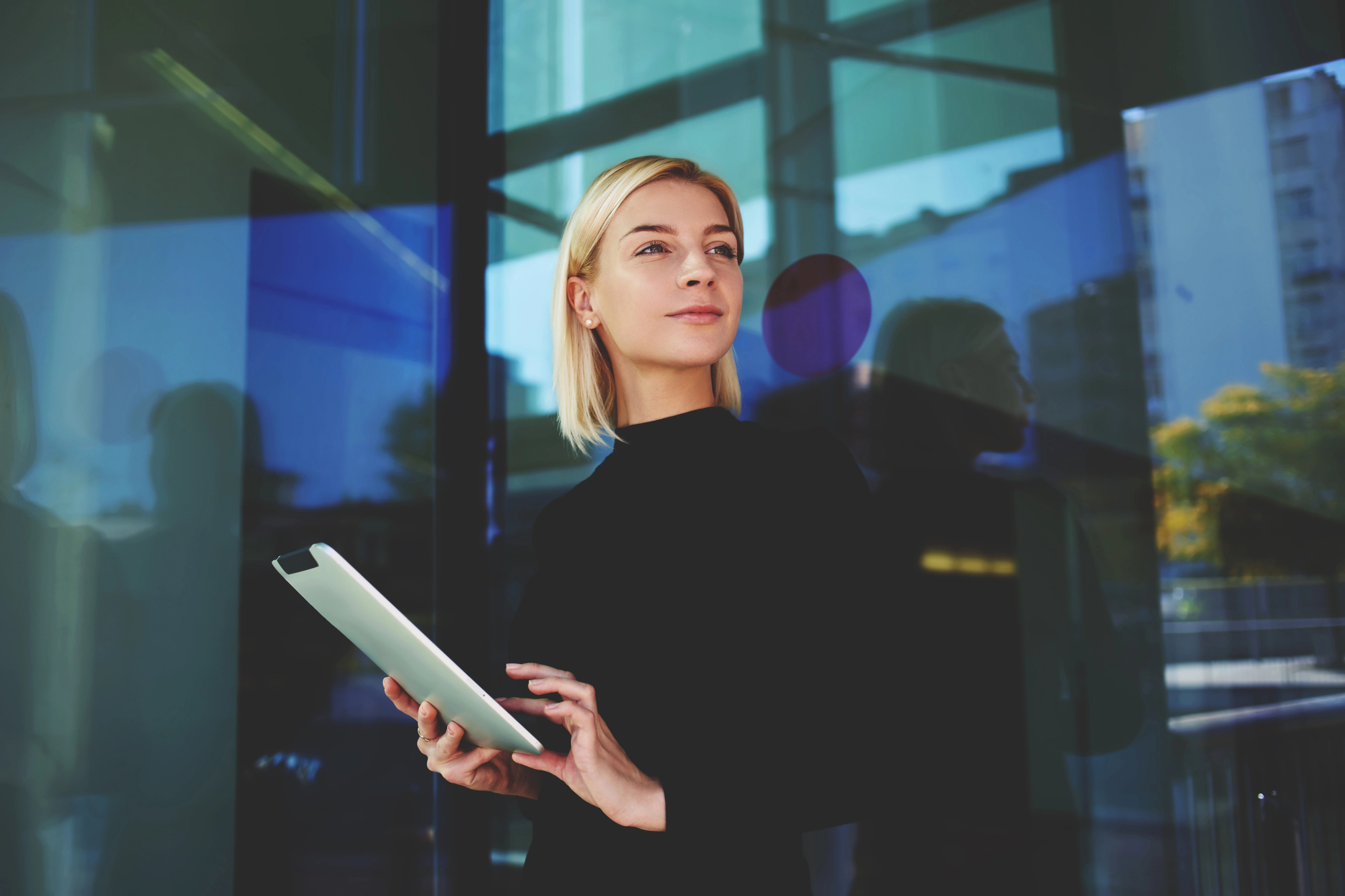 A woman in a black outfit stands confidently in front of a modern glass building, holding a tablet. The reflection of the cityscape and trees is visible in the background.