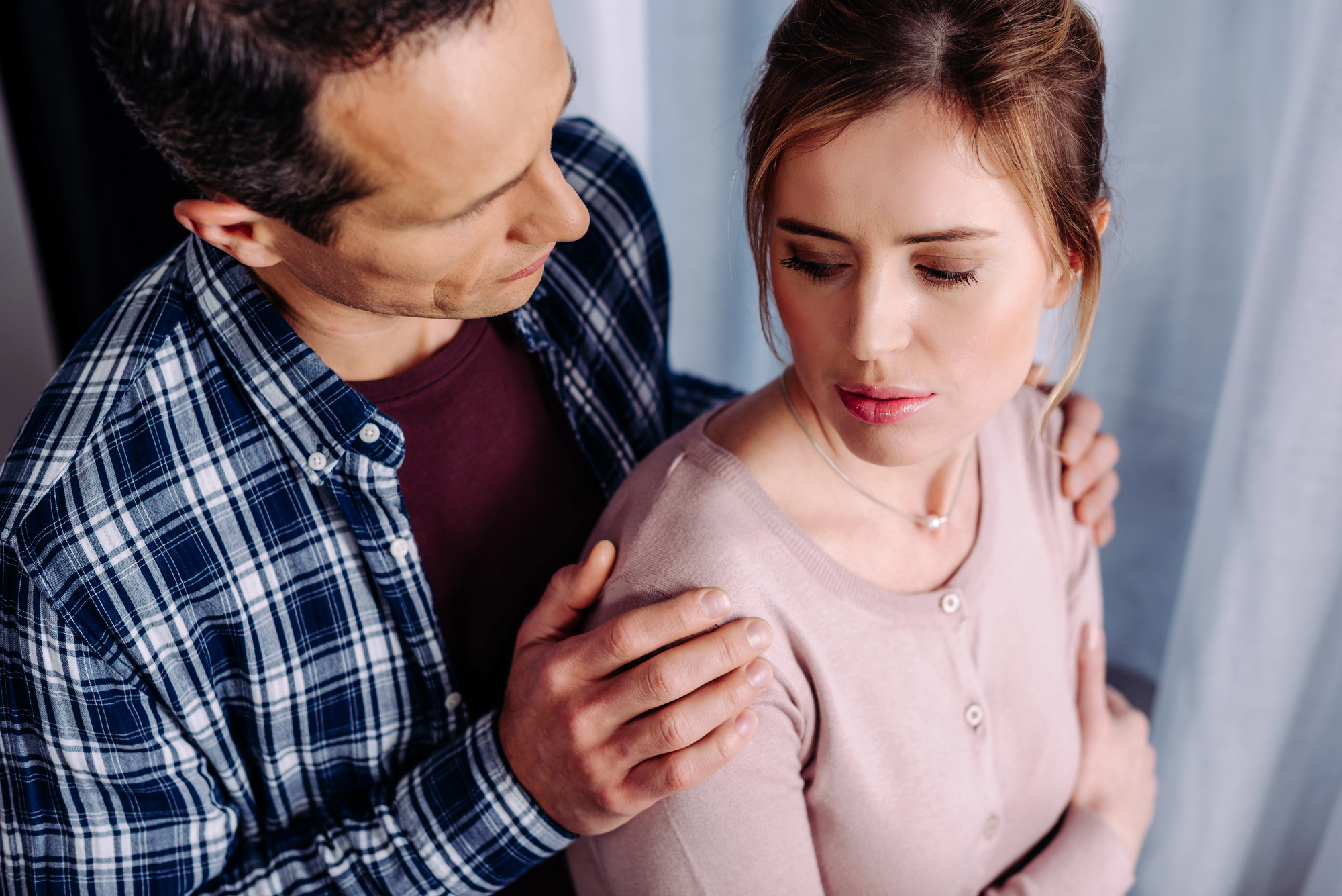 A man in a plaid shirt gently places his hands on the shoulders of a woman in a pink sweater. The woman looks away, appearing thoughtful. They are indoors with a curtain in the background.