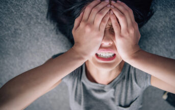 A person lying down with their hands covering their face, appearing distressed. They are wearing a gray shirt and have dark hair spread out on a gray surface.