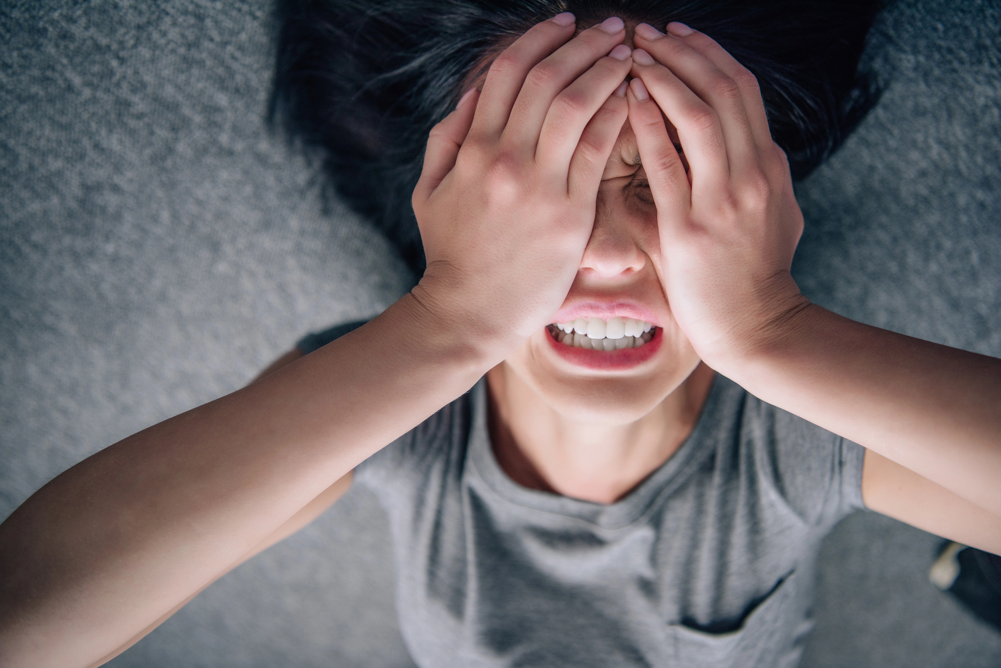 A person lying down with their hands covering their face, appearing distressed. They are wearing a gray shirt and have dark hair spread out on a gray surface.