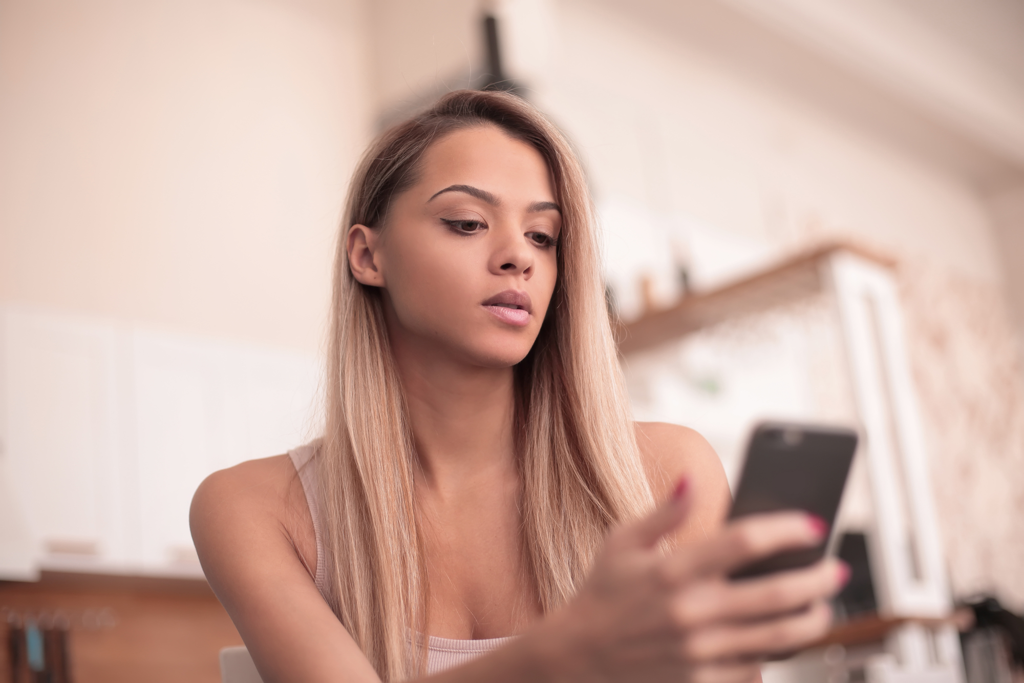 A woman with long blonde hair, wearing a light-colored tank top, sits indoors looking at her smartphone. The background features a blurred view of white cabinets and a countertop, indicating she is in a kitchen or dining area.