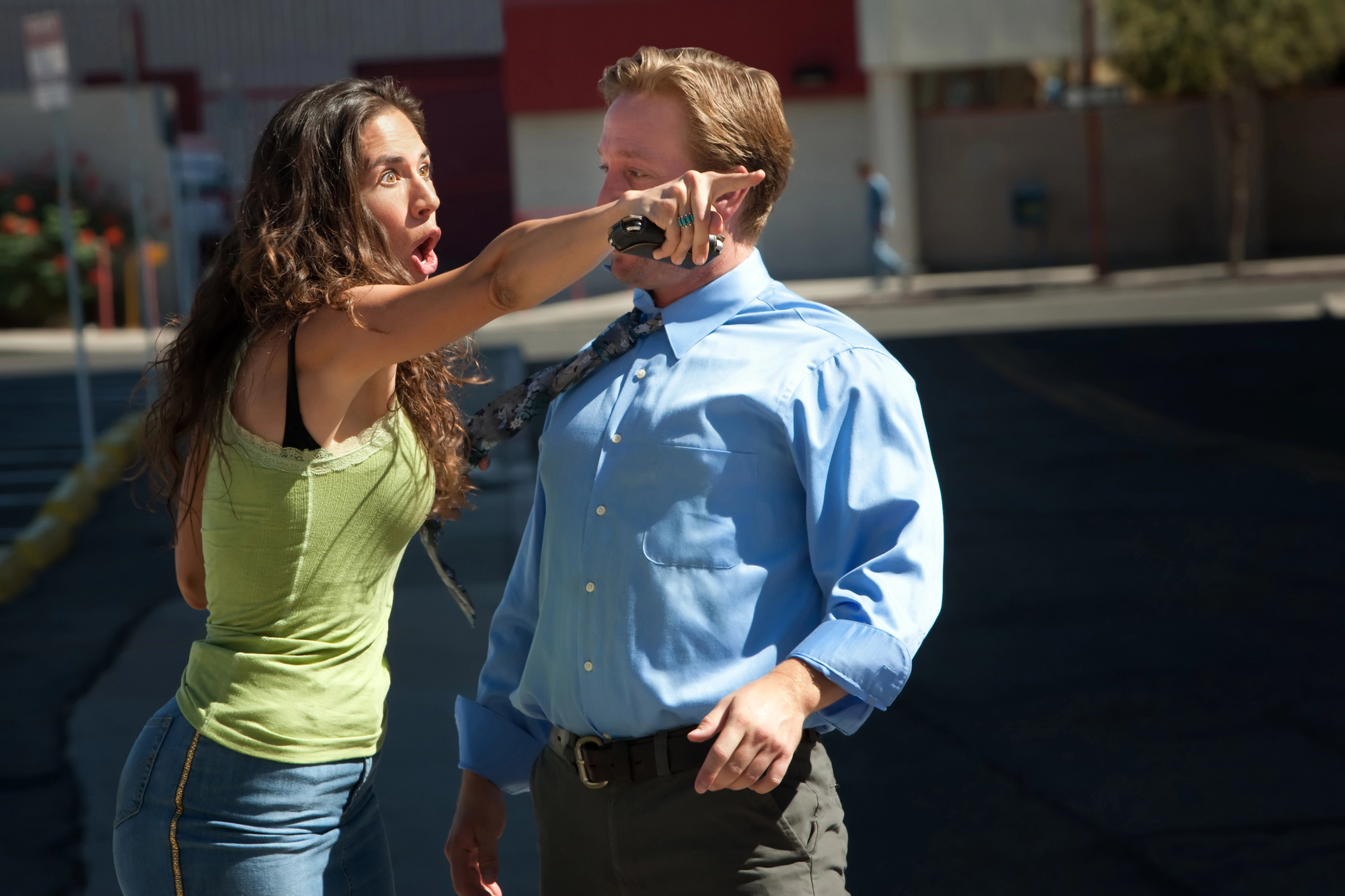 A woman with long hair angrily points in the distance while holding a phone. A man in a blue shirt stands beside her, looking in the same direction. They appear to be in an urban setting with buildings in the background.