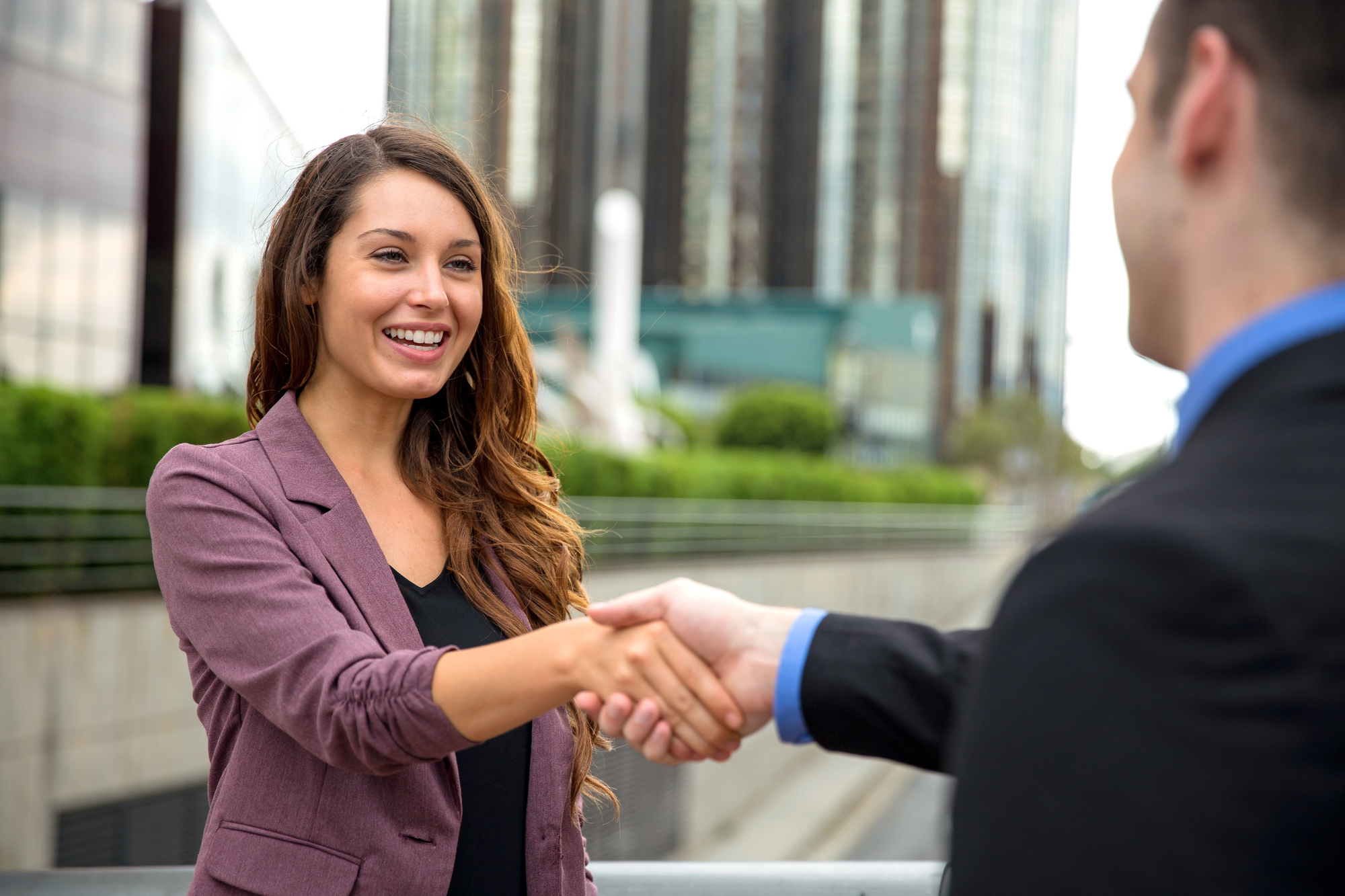 A woman in a purple blazer smiles as she shakes hands with a man in a suit. They stand outdoors in a city setting, with tall buildings and greenery in the background.