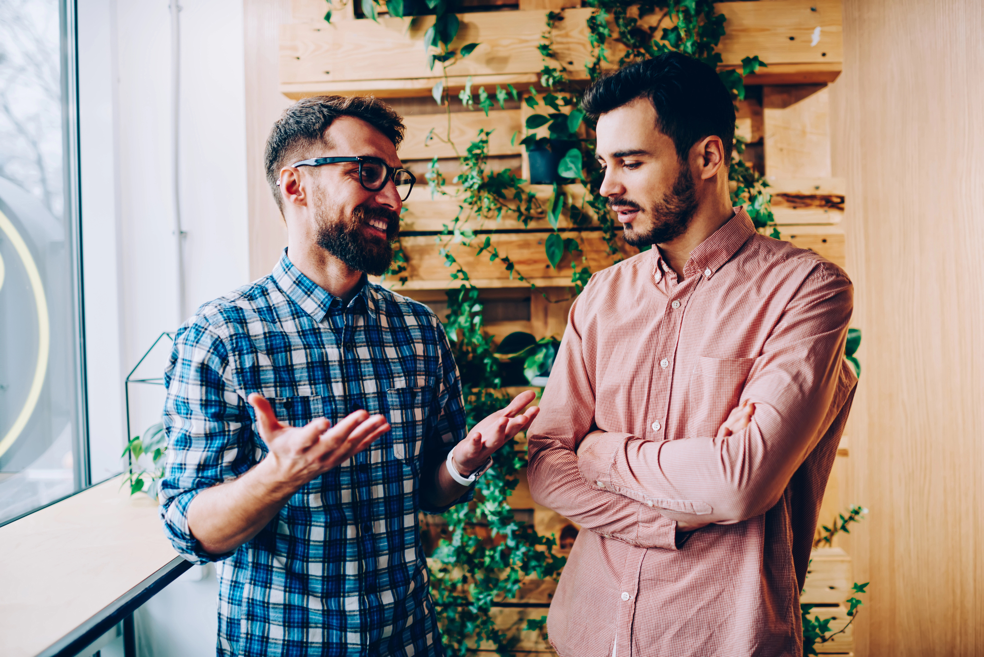 Two men are having a conversation in a bright room. One is gesturing with his hands, wearing a plaid shirt, and glasses. The other has his arms crossed, wearing a pink shirt. Behind them is a wooden wall with green plants.