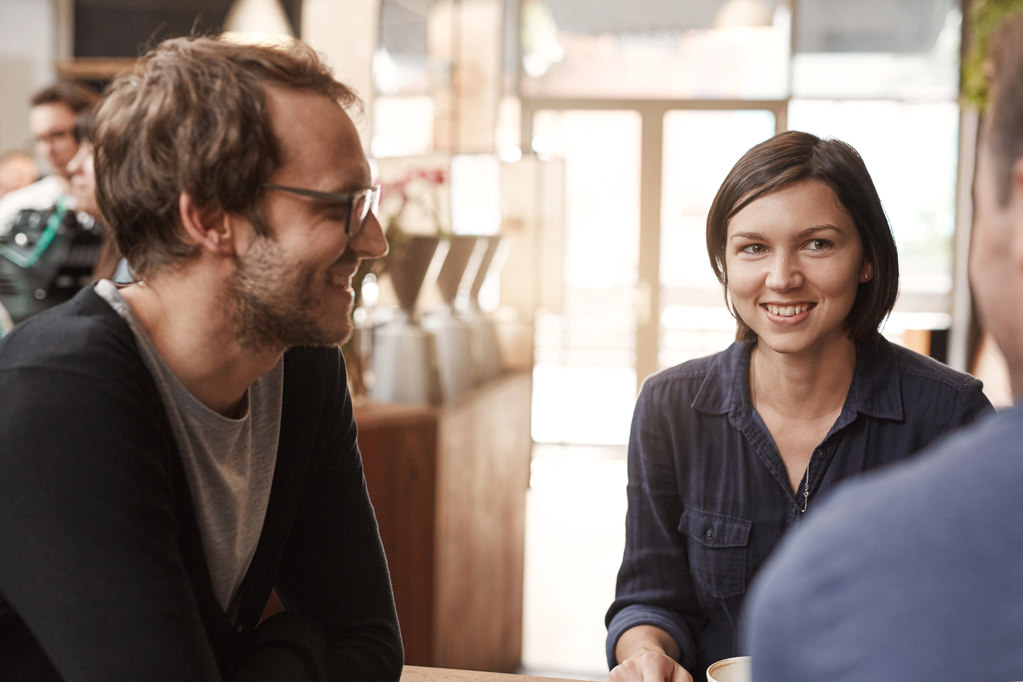 Three people are sitting and conversing at a table in a cozy café. A man with glasses and a beard on the left is smiling at a woman with short dark hair in the center, who is also smiling. Another person is partially visible on the right, also engaged in the conversation.