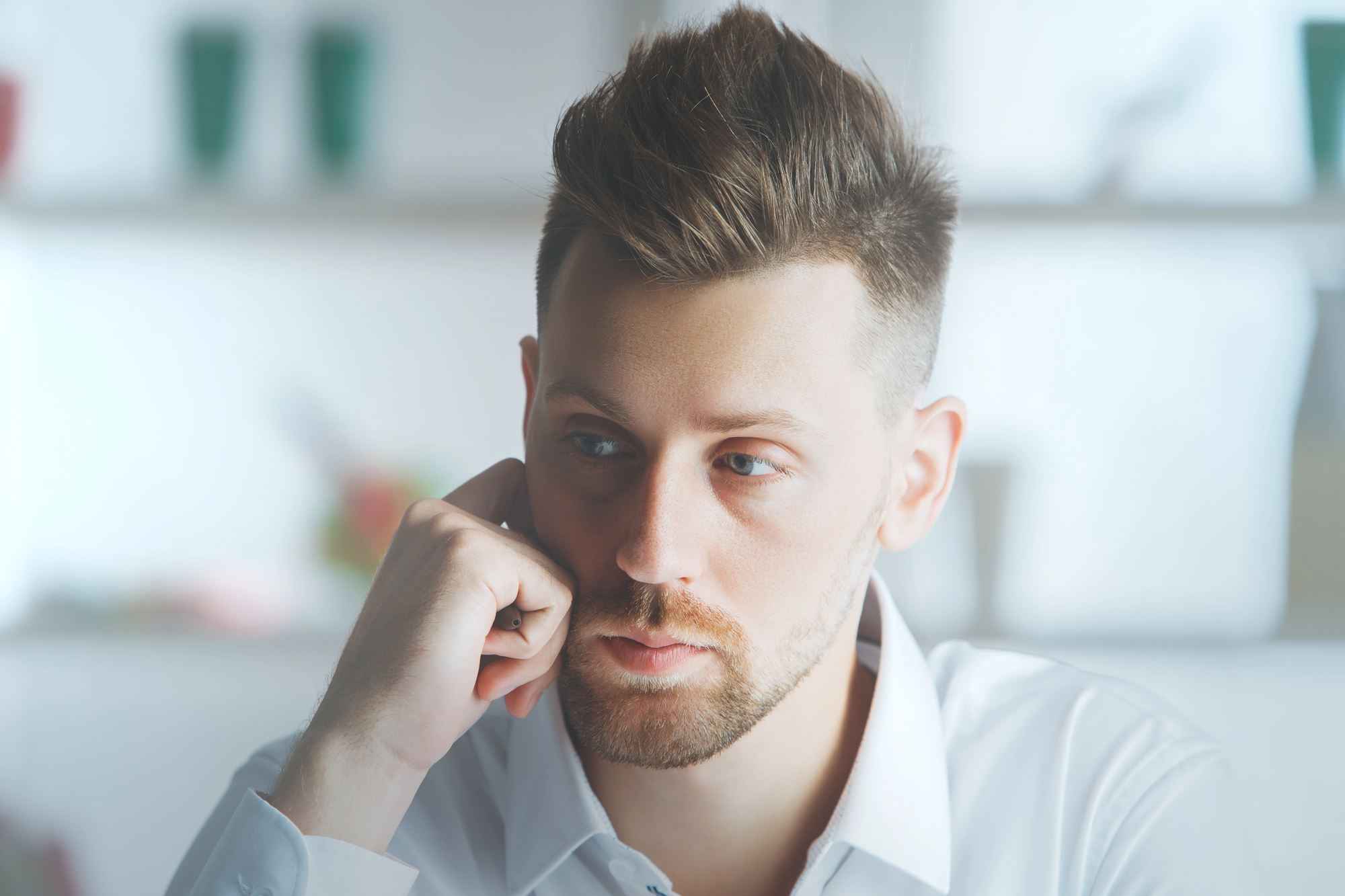 A person with short, styled hair and a trimmed beard rests their chin on a hand, gazing thoughtfully to the side. They are wearing a white shirt, and the background is softly blurred.