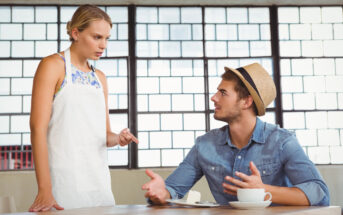 A woman in a white apron gestures with her finger at a man sitting at a table. The man, wearing a hat and denim shirt, looks up at her while gesturing with his hands. They are in a modern room with large grid windows. A coffee cup is on the table.