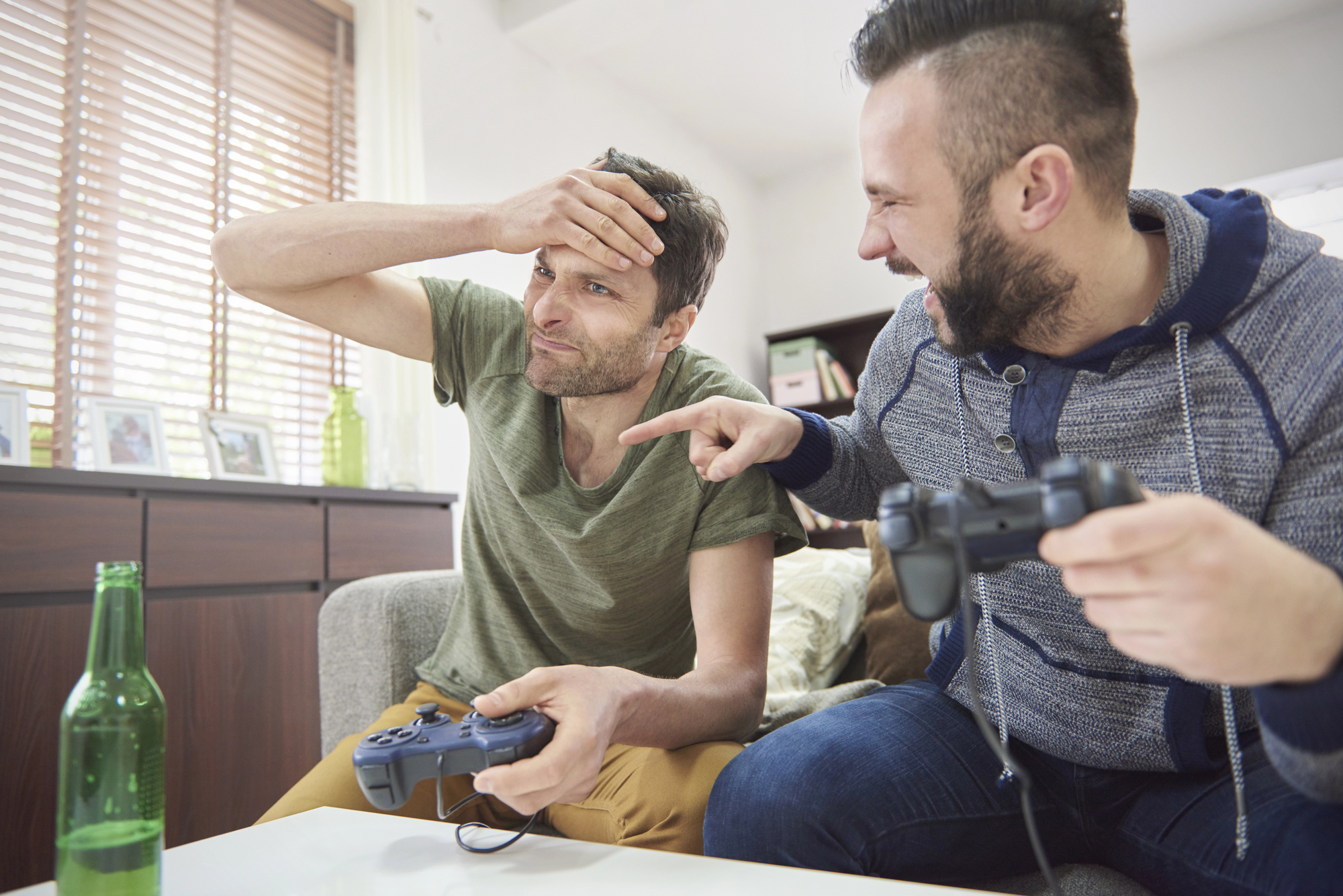 Two men sitting on a couch playing video games. The man on the left looks frustrated, holding his forehead, while the man on the right laughs and points at him. A gaming controller is in each of their hands, and a green bottle is on the table.