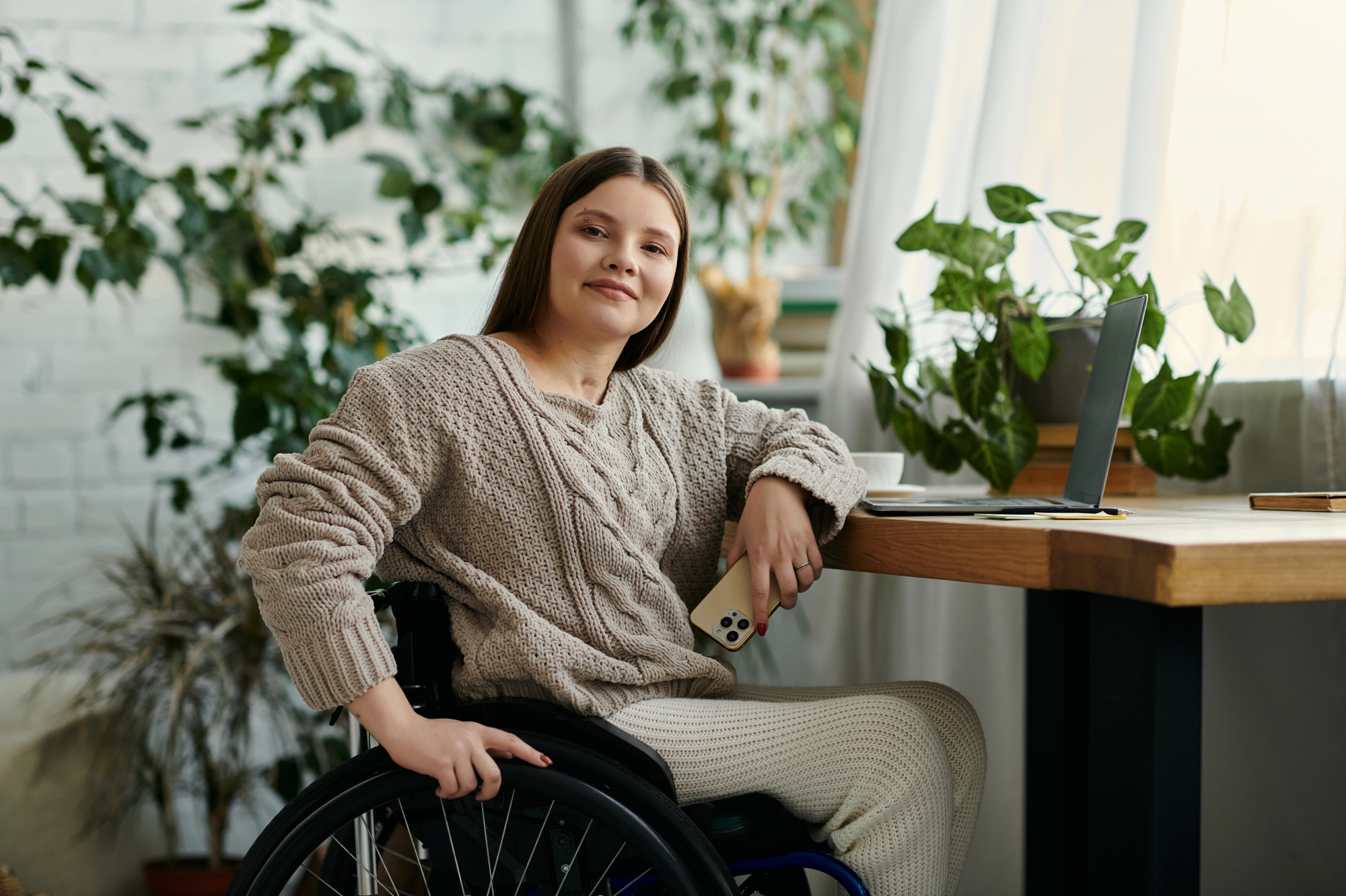 A woman in a wheelchair sits at a table with a laptop, holding a smartphone. She wears a cozy sweater and pants, smiling in a room filled with green plants and soft natural light.