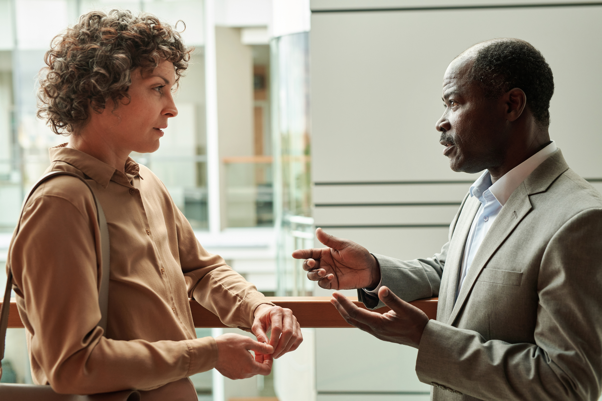 Two people in a serious conversation inside a modern building. The person on the left wears a brown shirt, and the person on the right is in a gray suit. They are standing by a railing, engaged in discussion.