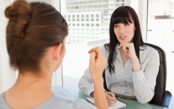 Two women are sitting in an office setting engaged in conversation. One woman, with dark hair, is listening attentively with her hand on her chin. The other woman, with her back to the camera, gestures while speaking. Both are seated at a glass table.