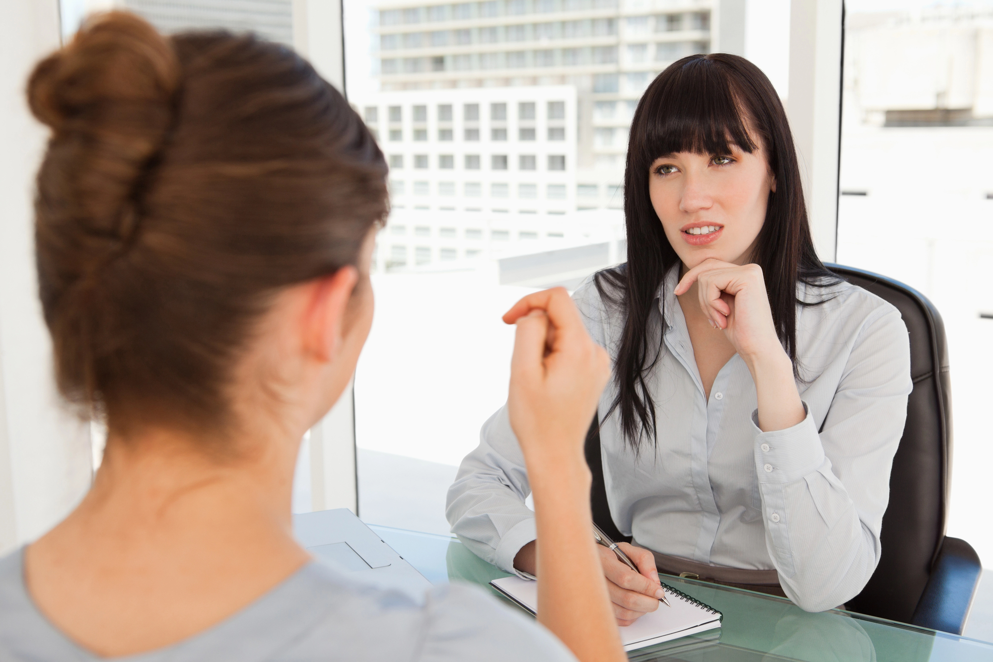 Two women are sitting in an office setting engaged in conversation. One woman, with dark hair, is listening attentively with her hand on her chin. The other woman, with her back to the camera, gestures while speaking. Both are seated at a glass table.