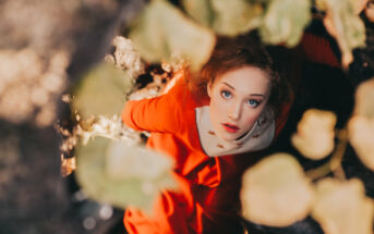 A woman with reddish-brown hair wearing an orange dress looks up through an opening surrounded by leaves. Sunlight partially illuminates her face and the leaves above her. The image is taken from a high angle, giving a sense of depth and nature.