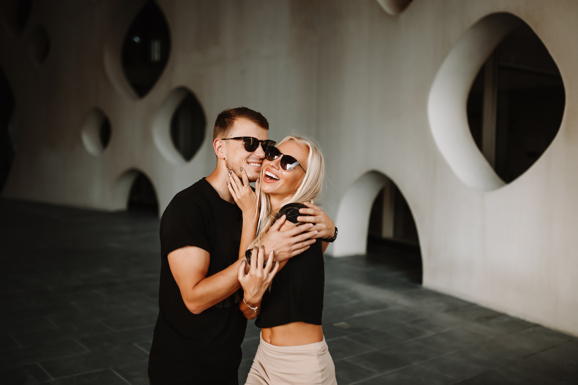 A couple wearing sunglasses embraces and laughs outdoors in front of a modern building with large geometric cutouts. They both have light skin and are dressed casually, with the man in a black t-shirt and the woman in a black top and beige pants.