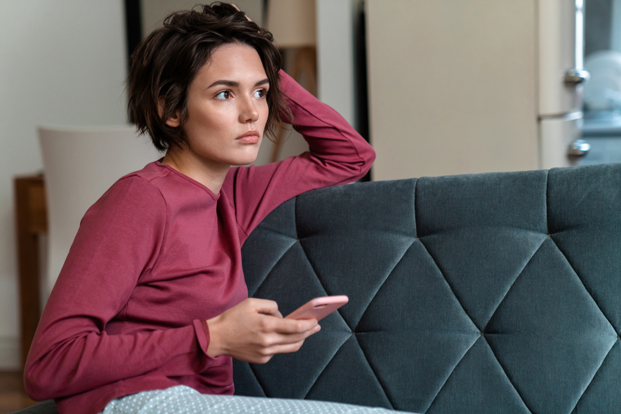 A woman with short brown hair, wearing a long-sleeved pink shirt, sits on a gray sofa holding a smartphone. She rests her head on her hand and looks thoughtfully into the distance. The room is softly lit.