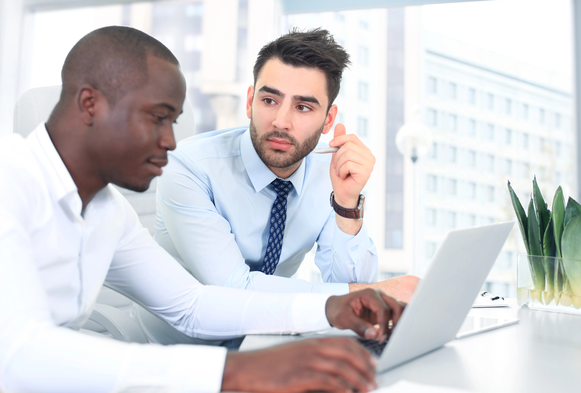 Two men are seated at a desk in an office, working on a laptop. One man is typing while the other listens attentively. They are dressed in business attire. A window with a cityscape is visible in the background, along with a small plant.