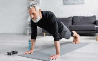 An older man with gray hair and beard performs a plank exercise on a gray yoga mat in a living room. He wears a black shirt and gray pants. Nearby are a water bottle and dumbbells. A gray couch is in the background.