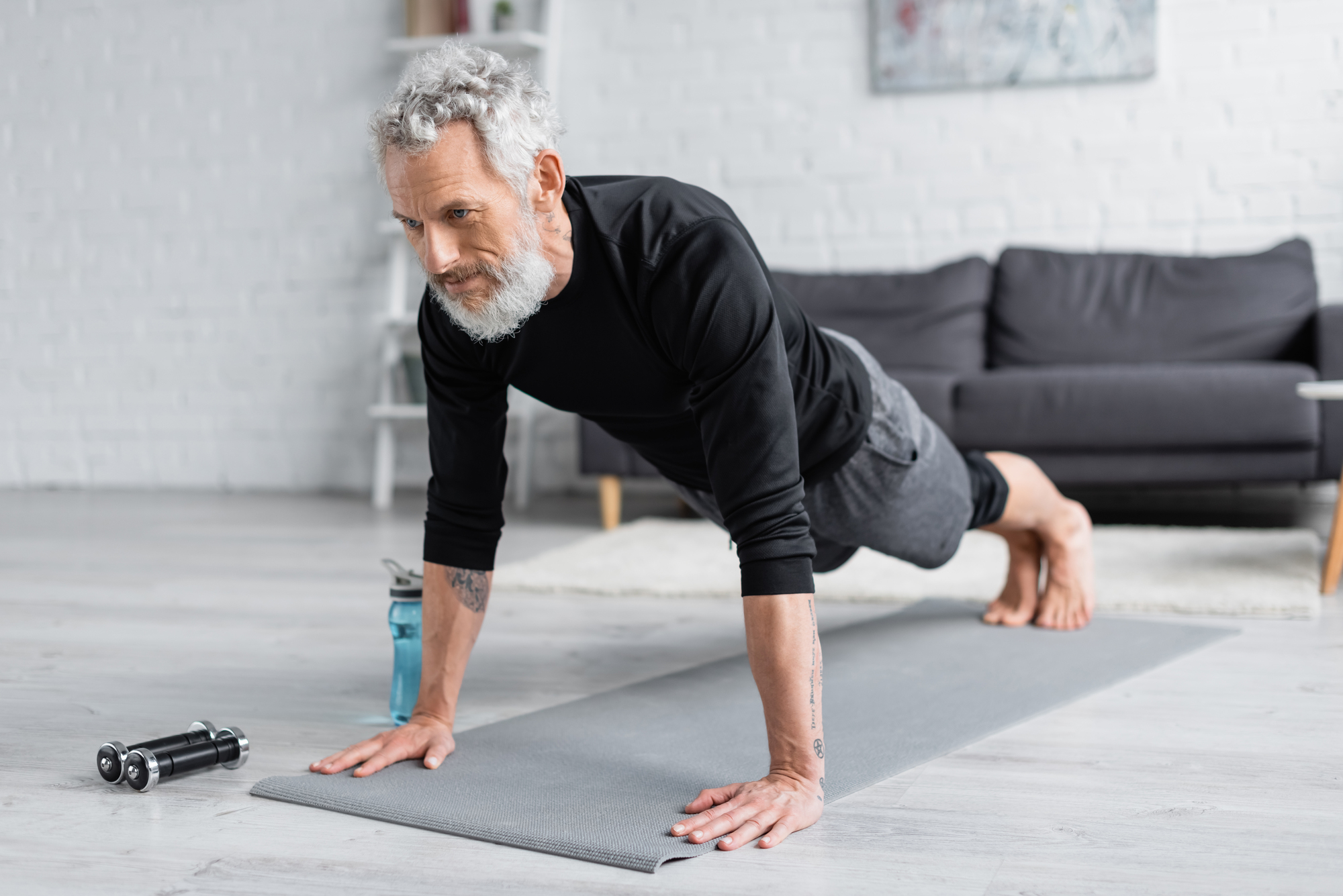 An older man with gray hair and beard performs a plank exercise on a gray yoga mat in a living room. He wears a black shirt and gray pants. Nearby are a water bottle and dumbbells. A gray couch is in the background.