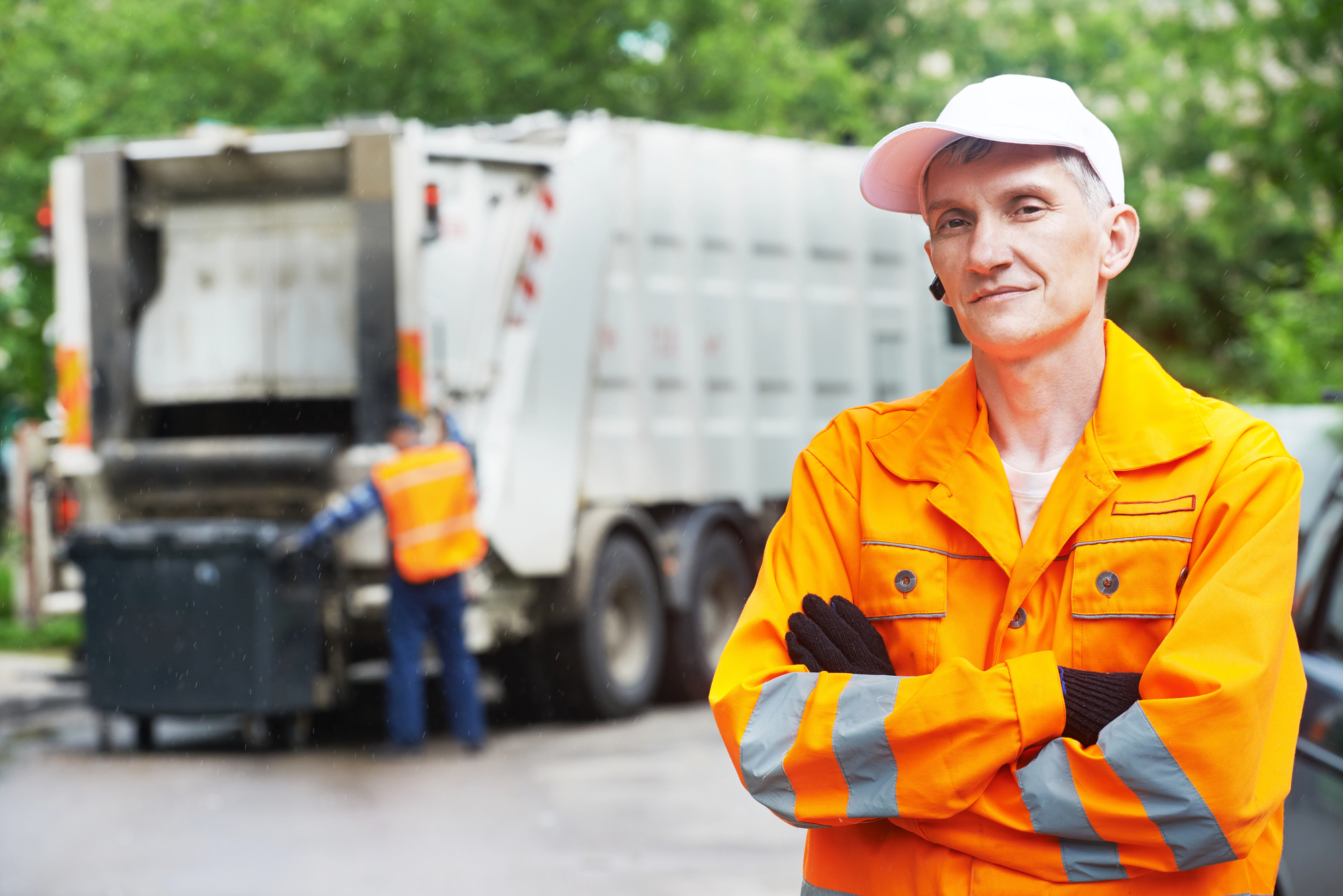 A sanitation worker in a bright orange uniform and white cap stands confidently with arms crossed in the foreground. A garbage truck is in the background, where another worker is operating the vehicle. Trees surround the scene.