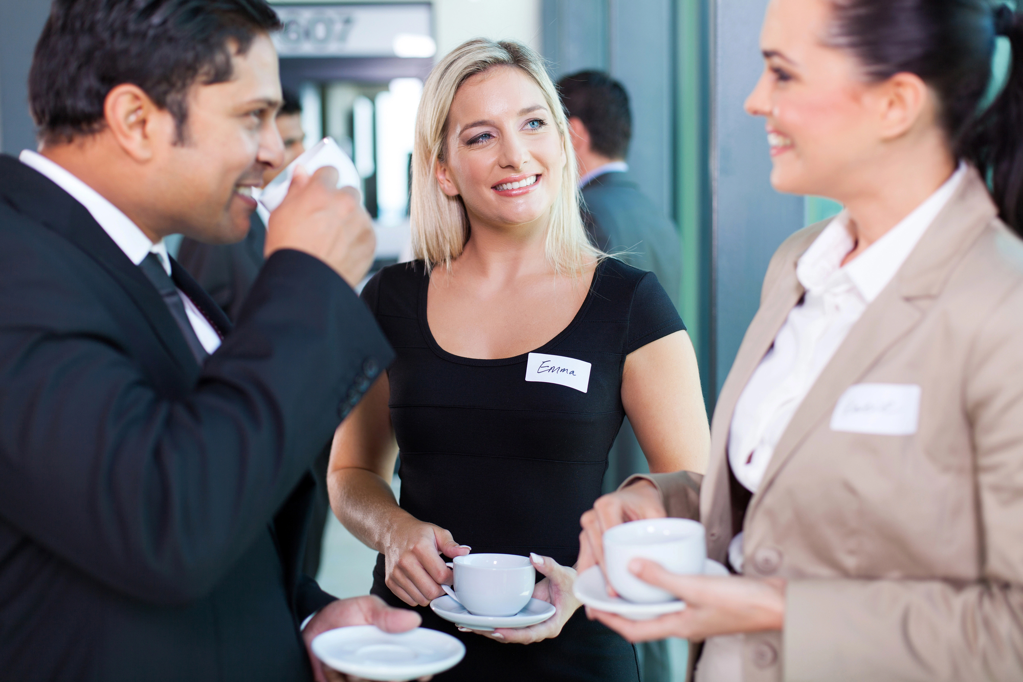 Three people wearing nametags are engaged in conversation while holding coffee cups. One man in a suit is sipping his drink, while two women are smiling. They appear to be at a professional event or conference.