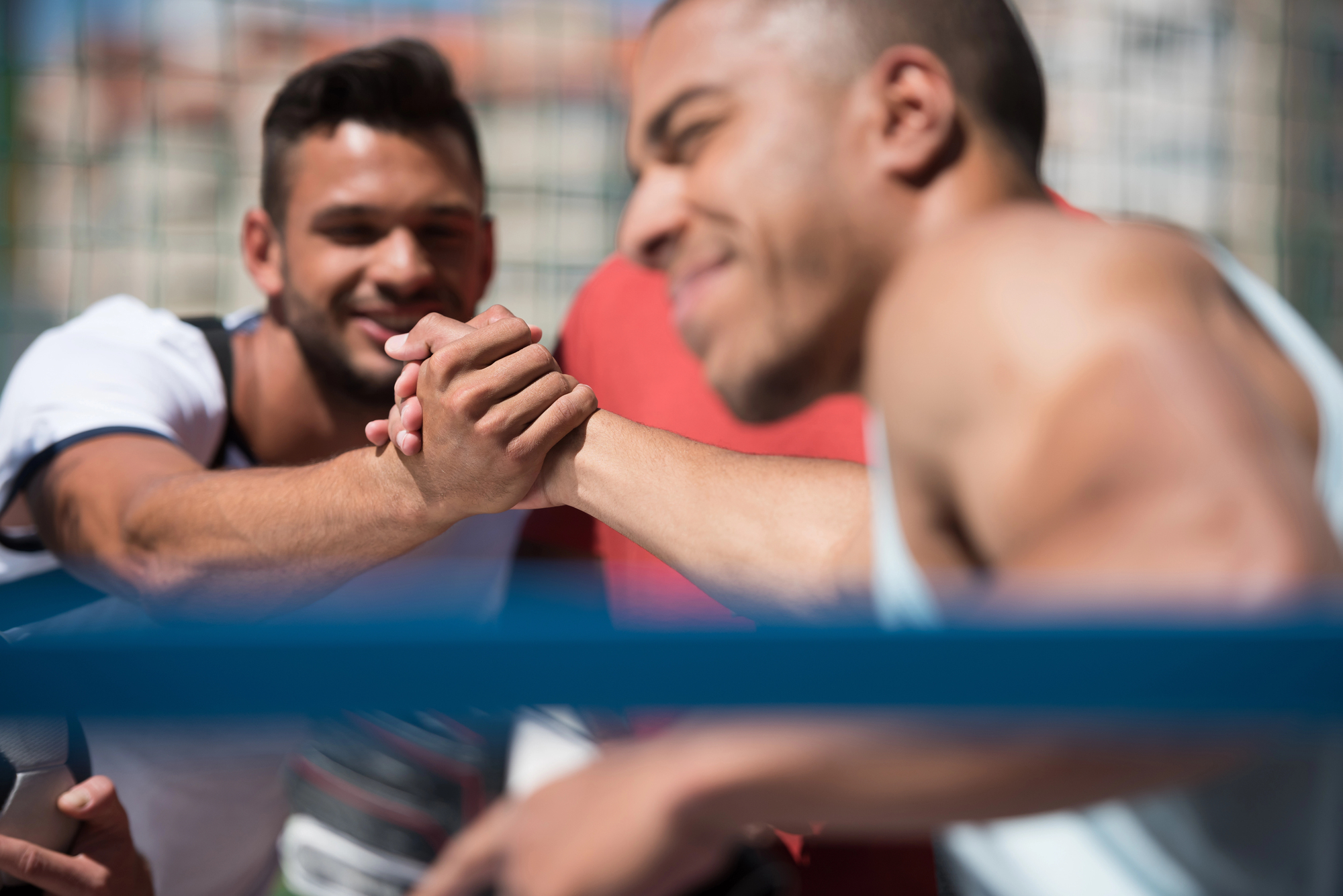 Two men arm wrestling outdoors, smiling as they compete with a blurred background. One is wearing a white shirt, the other a tank top. The scene conveys a friendly and energetic atmosphere.