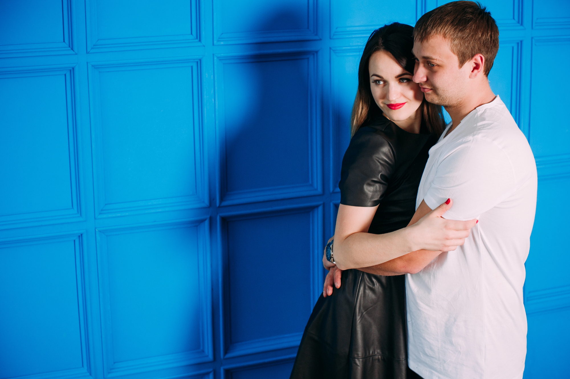 A woman in a black dress and a man in a white t-shirt embrace in front of a bright blue paneled wall. The woman is smiling while resting her head on the man's shoulder.