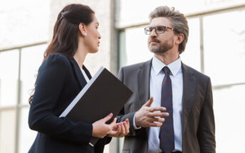 A man and a woman in business attire are having a conversation outdoors. The man, wearing glasses and a blue tie, gestures while speaking. The woman holds a black folder and listens attentively. They are in front of a glass building.