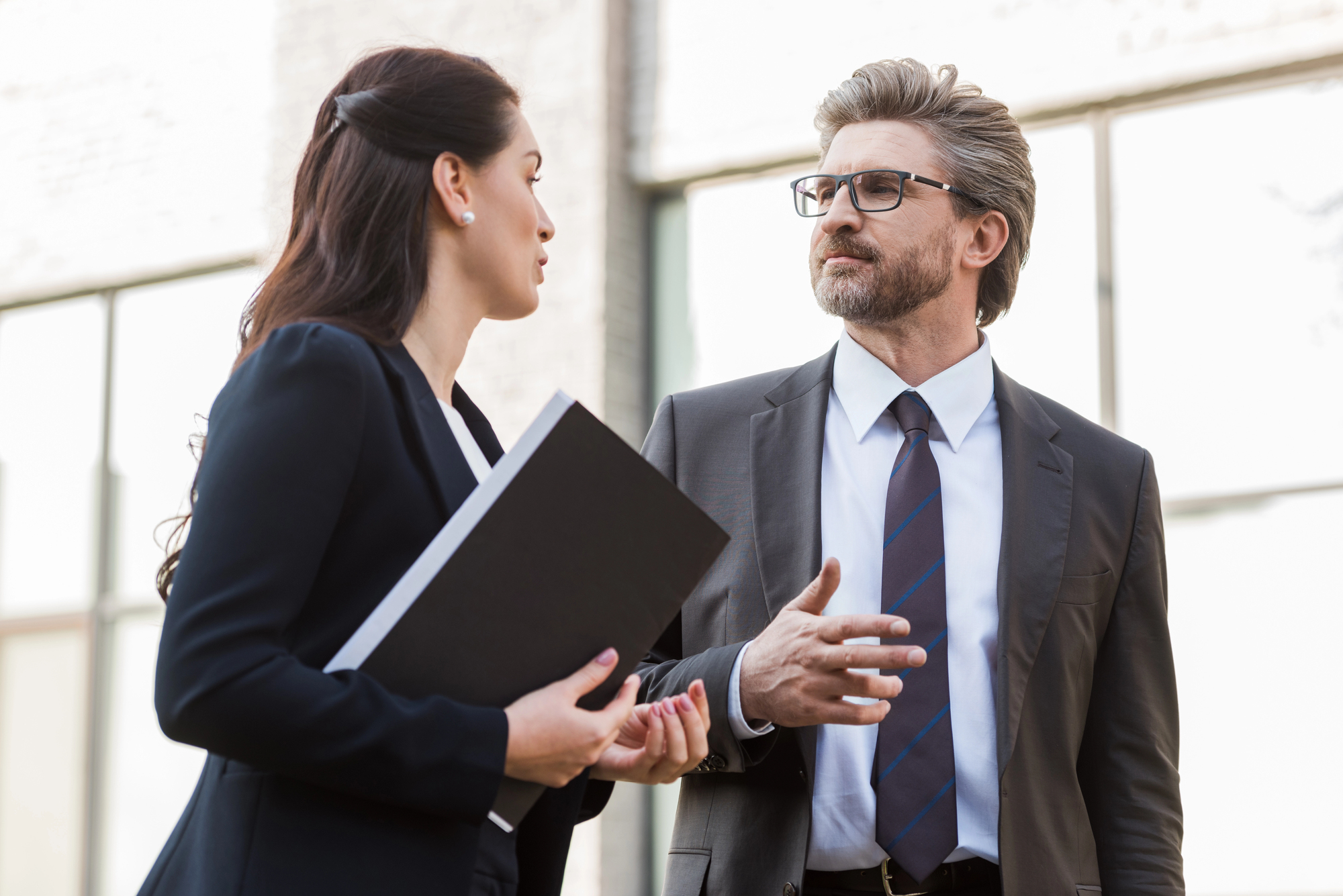 A man and a woman in business attire are having a conversation outdoors. The man, wearing glasses and a blue tie, gestures while speaking. The woman holds a black folder and listens attentively. They are in front of a glass building.