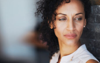 A woman with curly hair gazes thoughtfully to the side, leaning against a textured stone wall. She appears calm and contemplative, wearing a light-colored top. The background is softly blurred.