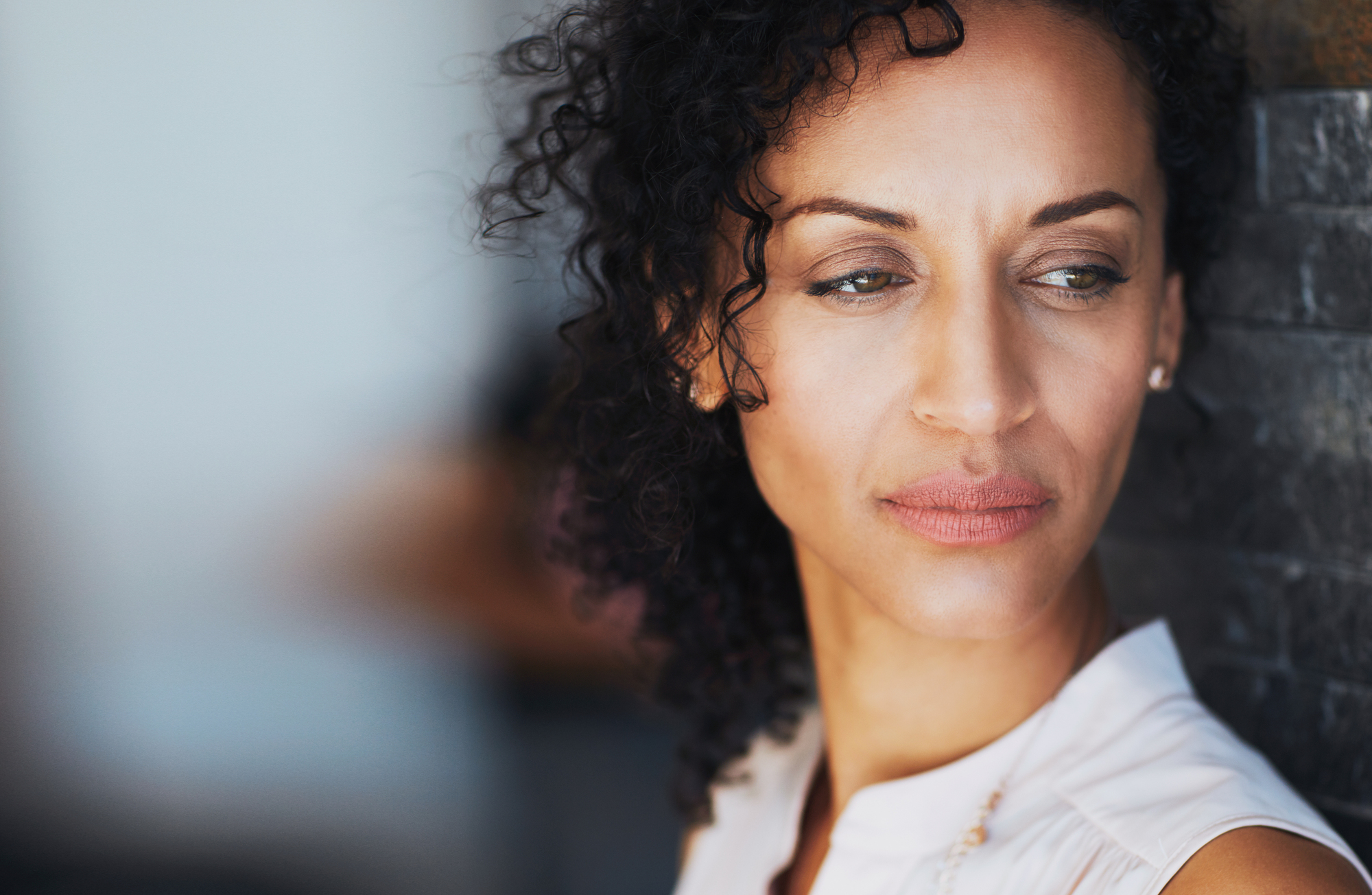 A woman with curly hair gazes thoughtfully to the side, leaning against a textured stone wall. She appears calm and contemplative, wearing a light-colored top. The background is softly blurred.