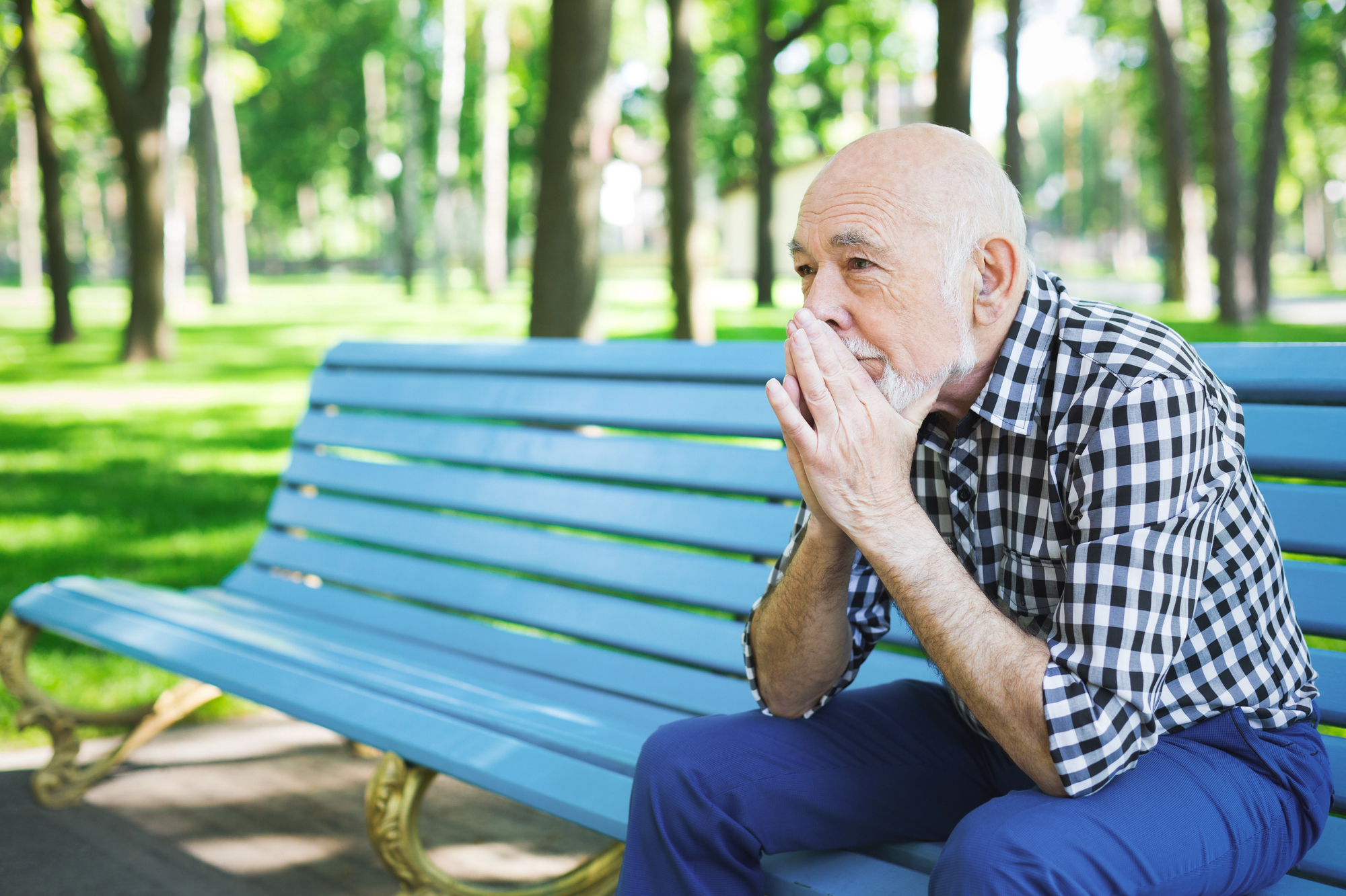 An elderly man with a beard, wearing a checkered shirt and blue pants, sits pensively on a blue bench in a sunlit park, surrounded by green trees. His hands cover his mouth, suggesting deep thought or concern.