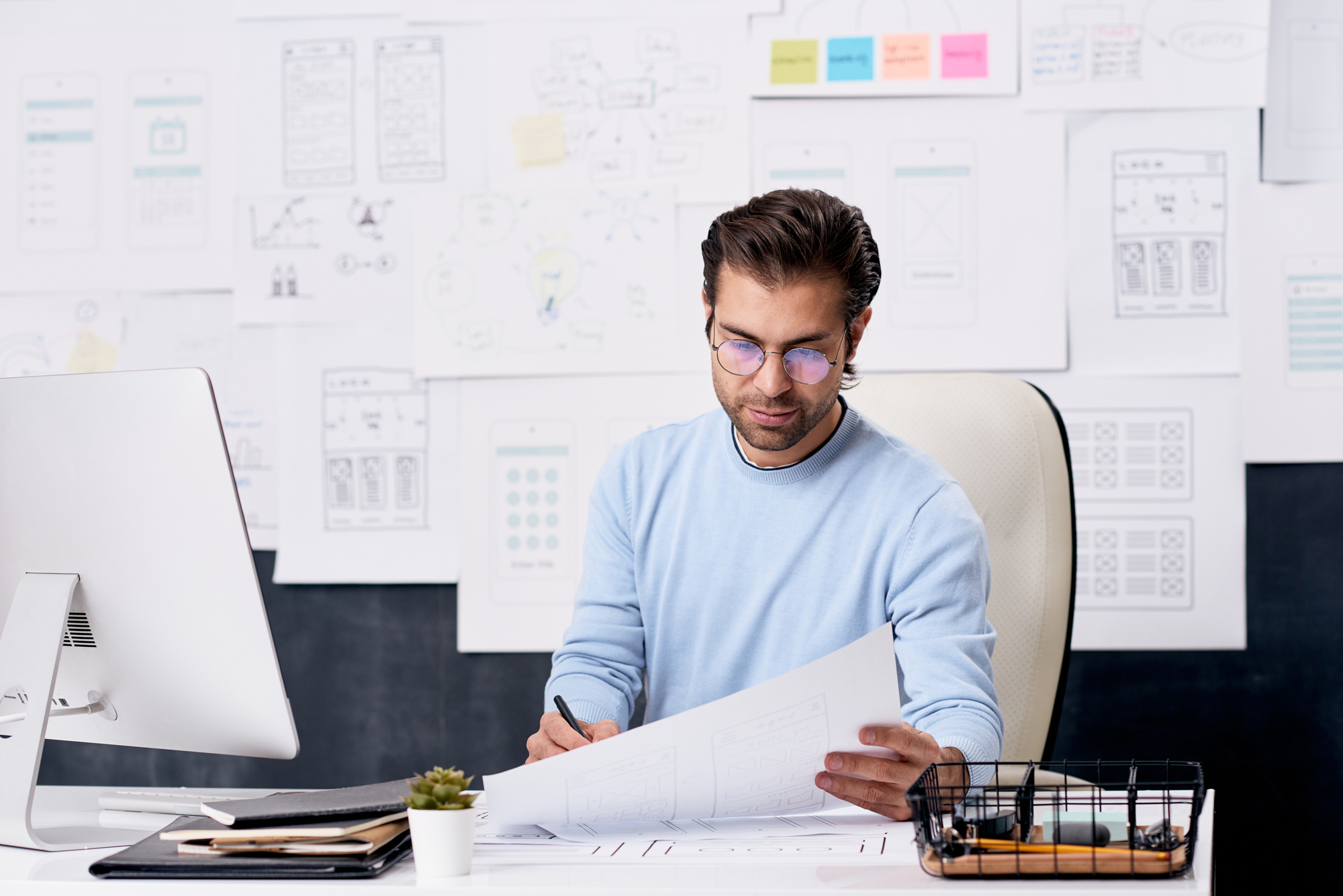 A man wearing glasses and a light blue sweater is seated at a desk, intently working on design drawings. The desk has a plant, a container with supplies, and a computer. The background features numerous design sketches pinned to the wall.