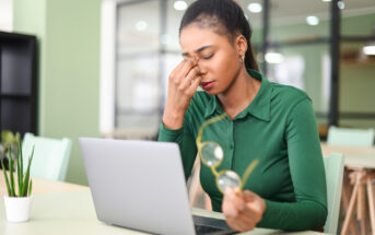 A woman in a green shirt sits at a desk with a laptop, holding her eyeglasses in one hand and rubbing her eyes with the other, appearing tired or stressed. A small plant is on the table nearby. The background features office furniture.