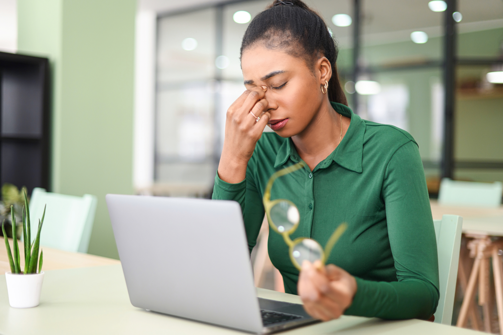 A woman in a green shirt sits at a desk with a laptop, holding her eyeglasses in one hand and rubbing her eyes with the other, appearing tired or stressed. A small plant is on the table nearby. The background features office furniture.