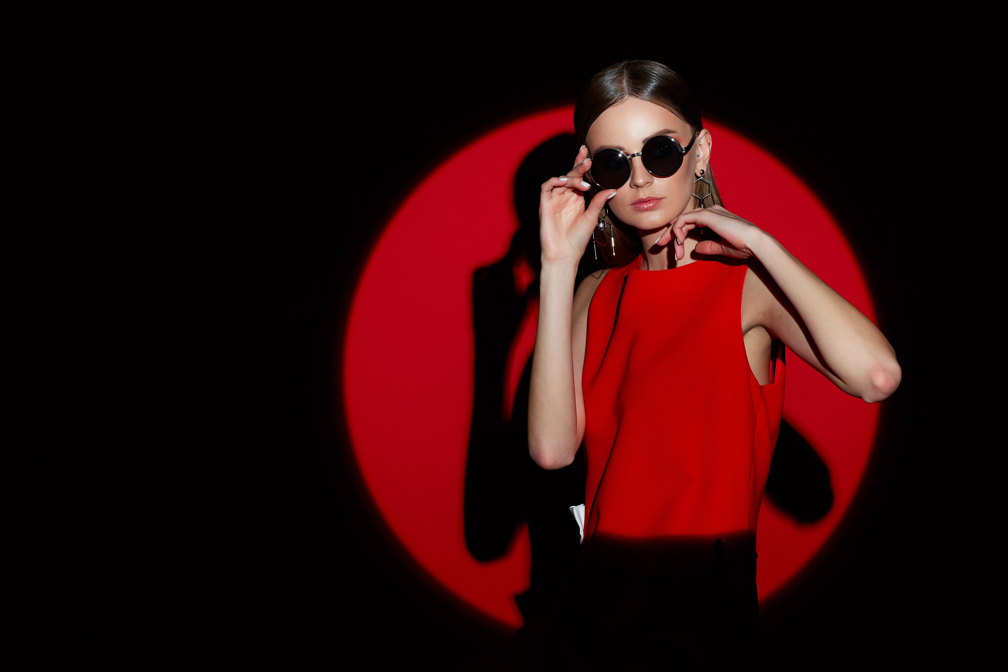 A woman in a red dress stands against a black background with a large red circle. She wears round sunglasses and adjusts them while looking at the camera. Her hair is sleek and she wears hoop earrings.