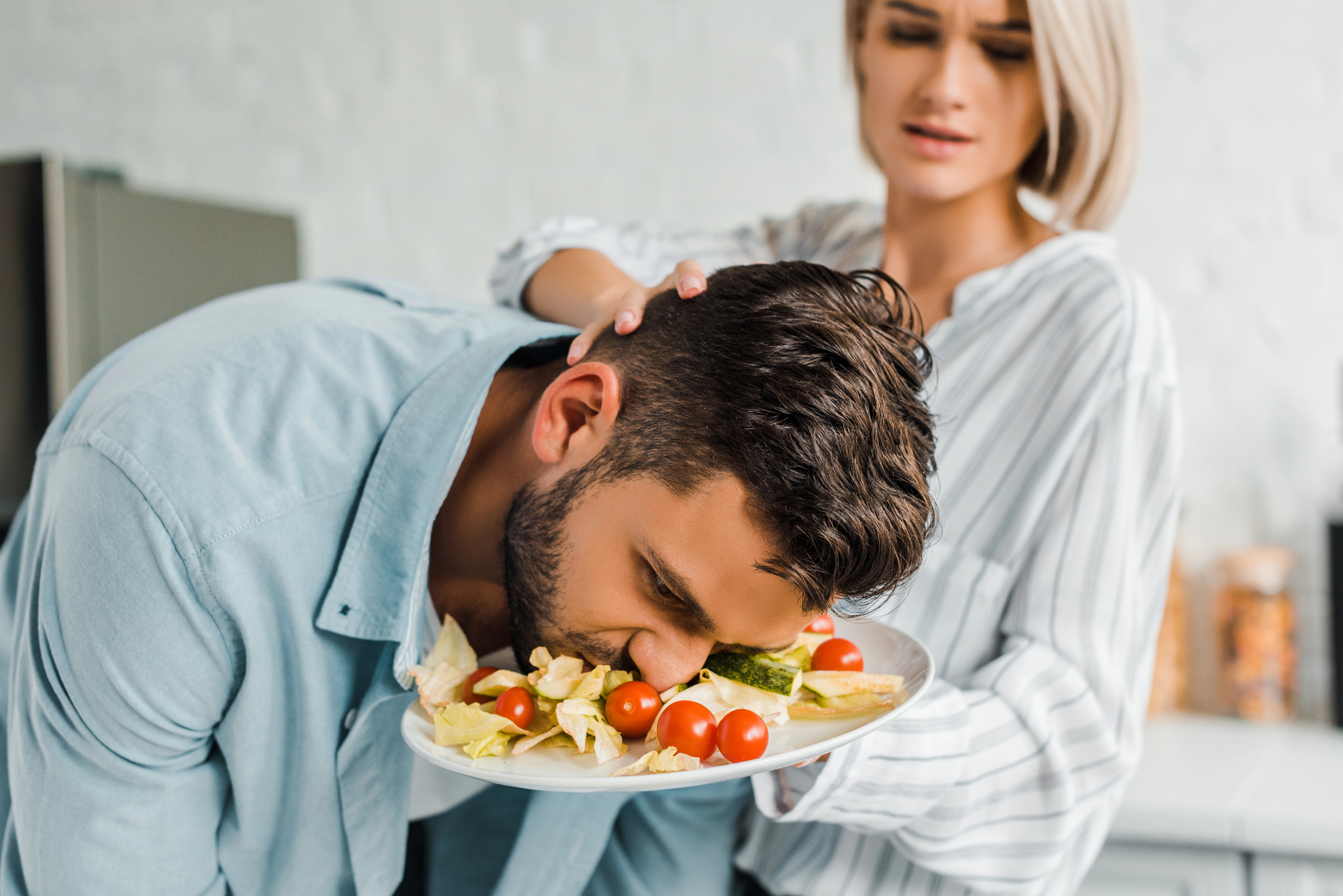 A man playfully dips his face into a plate of salad while a woman stands next to him with a surprised expression. The plate contains lettuce, cherry tomatoes, and sliced cucumbers. They are in a kitchen setting.