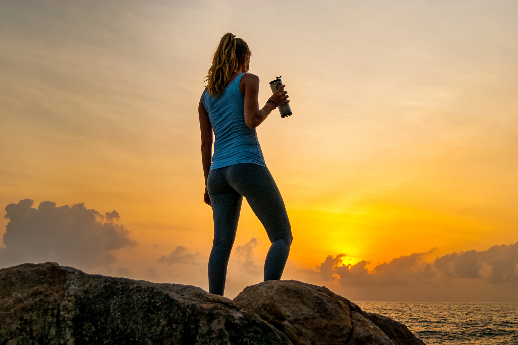 A woman stands on rocks by the sea, holding a water bottle and gazing at the vibrant sunset. She wears a blue tank top and gray leggings, with a ponytail. The sky is filled with warm hues of orange and yellow.