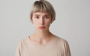 A person with short, light blonde hair and a neutral expression looks directly at the camera. They are wearing a light beige shirt, and the background is plain and light-colored.