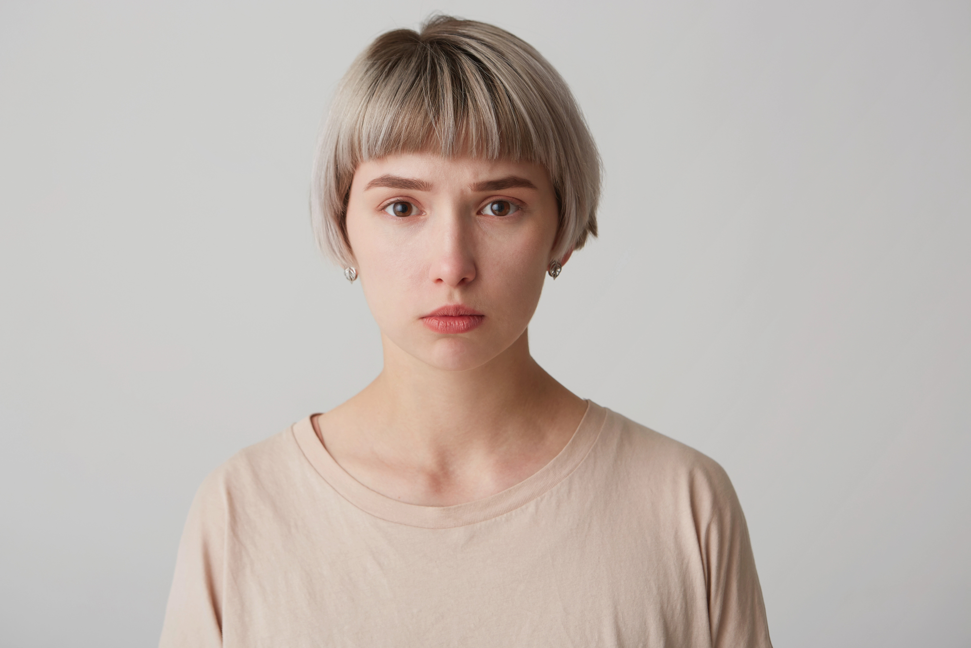 A person with short, light blonde hair and a neutral expression looks directly at the camera. They are wearing a light beige shirt, and the background is plain and light-colored.