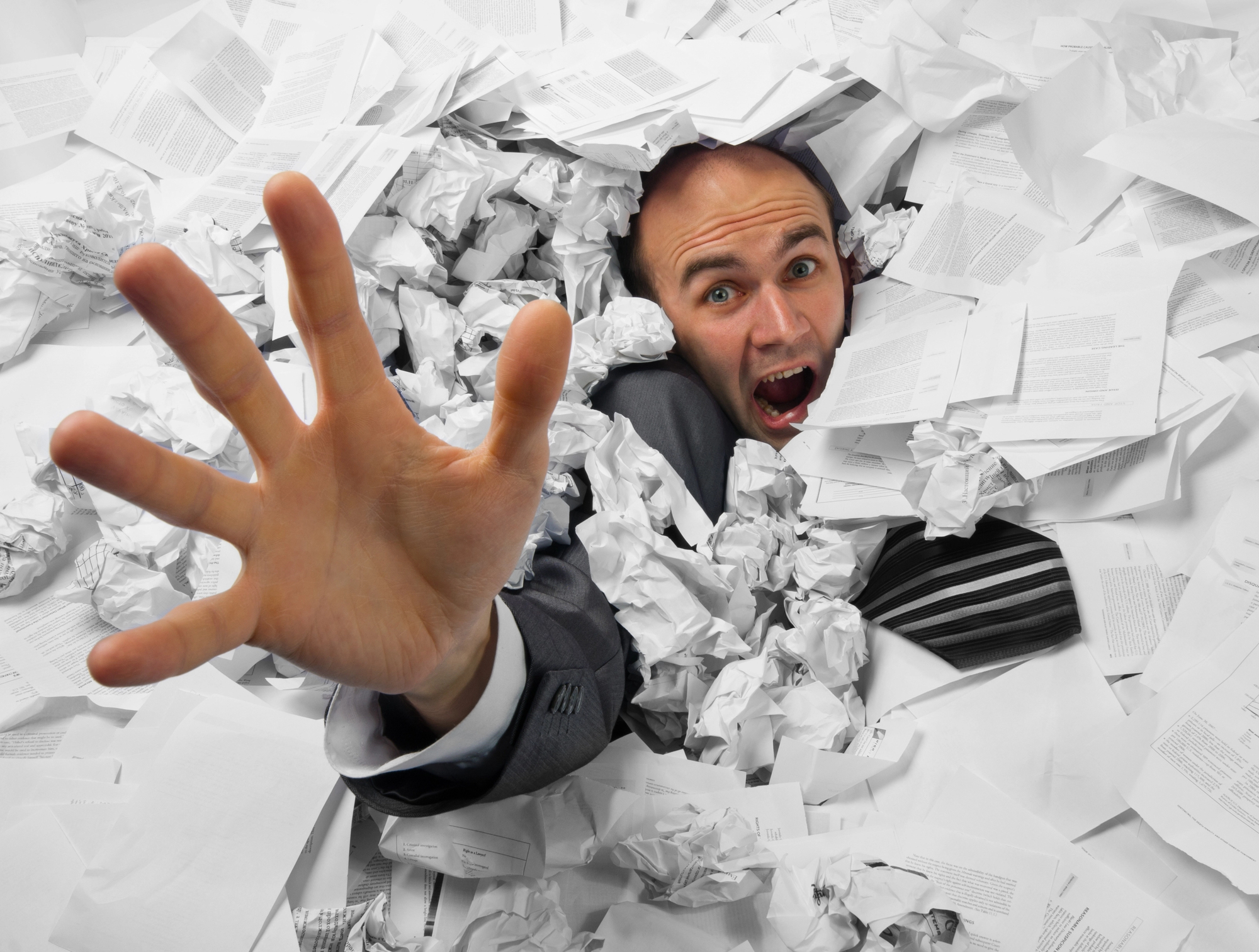 A man in a suit is overwhelmed by a large pile of crumpled papers, reaching out with a distressed expression. His hand is extended toward the camera as he appears to be buried in the disorganized documents.