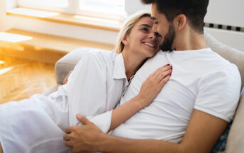 A couple is cuddling and smiling at each other on a couch in a sunlit room. The woman, wearing a white shirt, rests her head on the man's chest. The man, in a white T-shirt, holds her close, creating an atmosphere of warmth and affection.