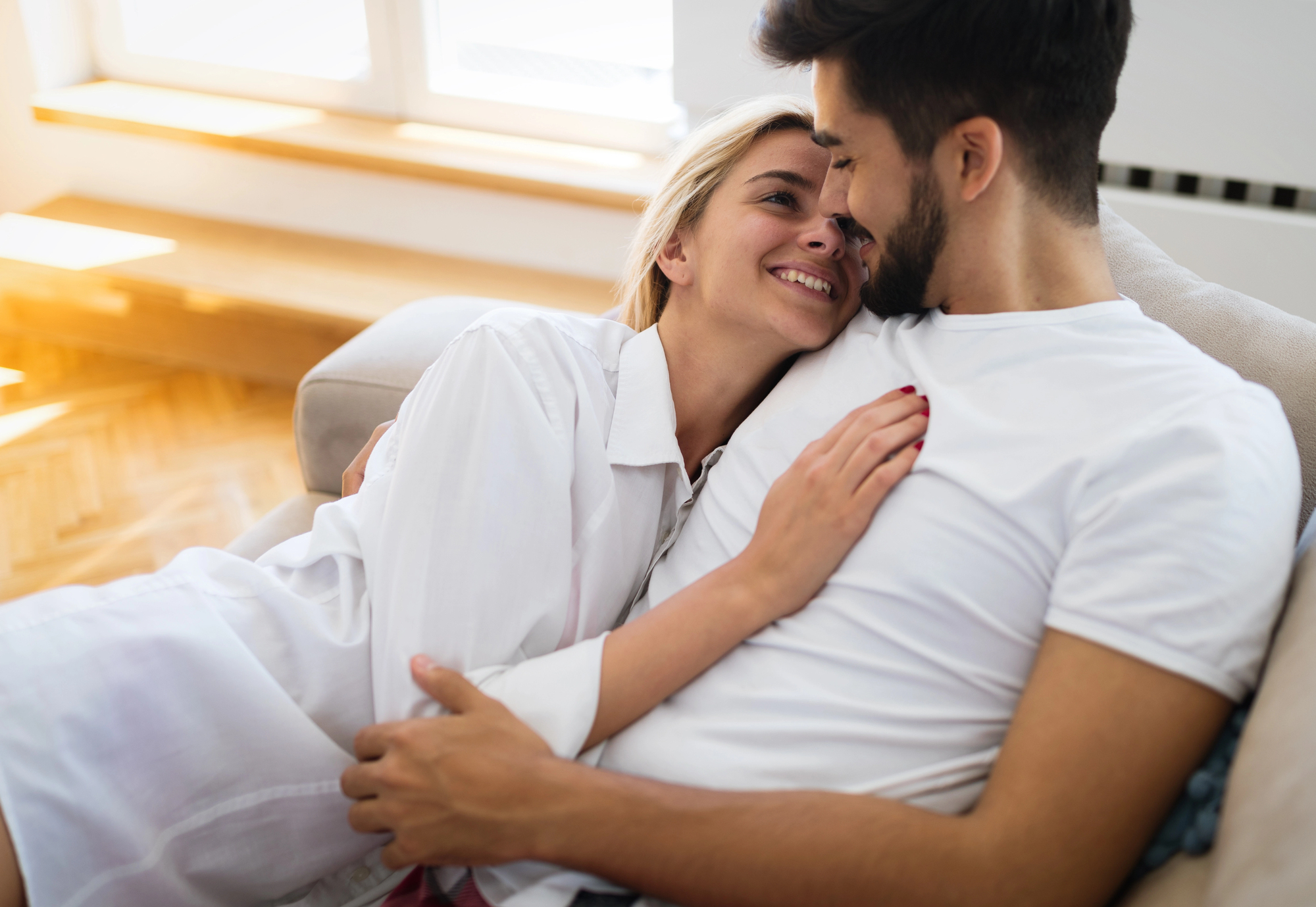 A couple is cuddling and smiling at each other on a couch in a sunlit room. The woman, wearing a white shirt, rests her head on the man's chest. The man, in a white T-shirt, holds her close, creating an atmosphere of warmth and affection.