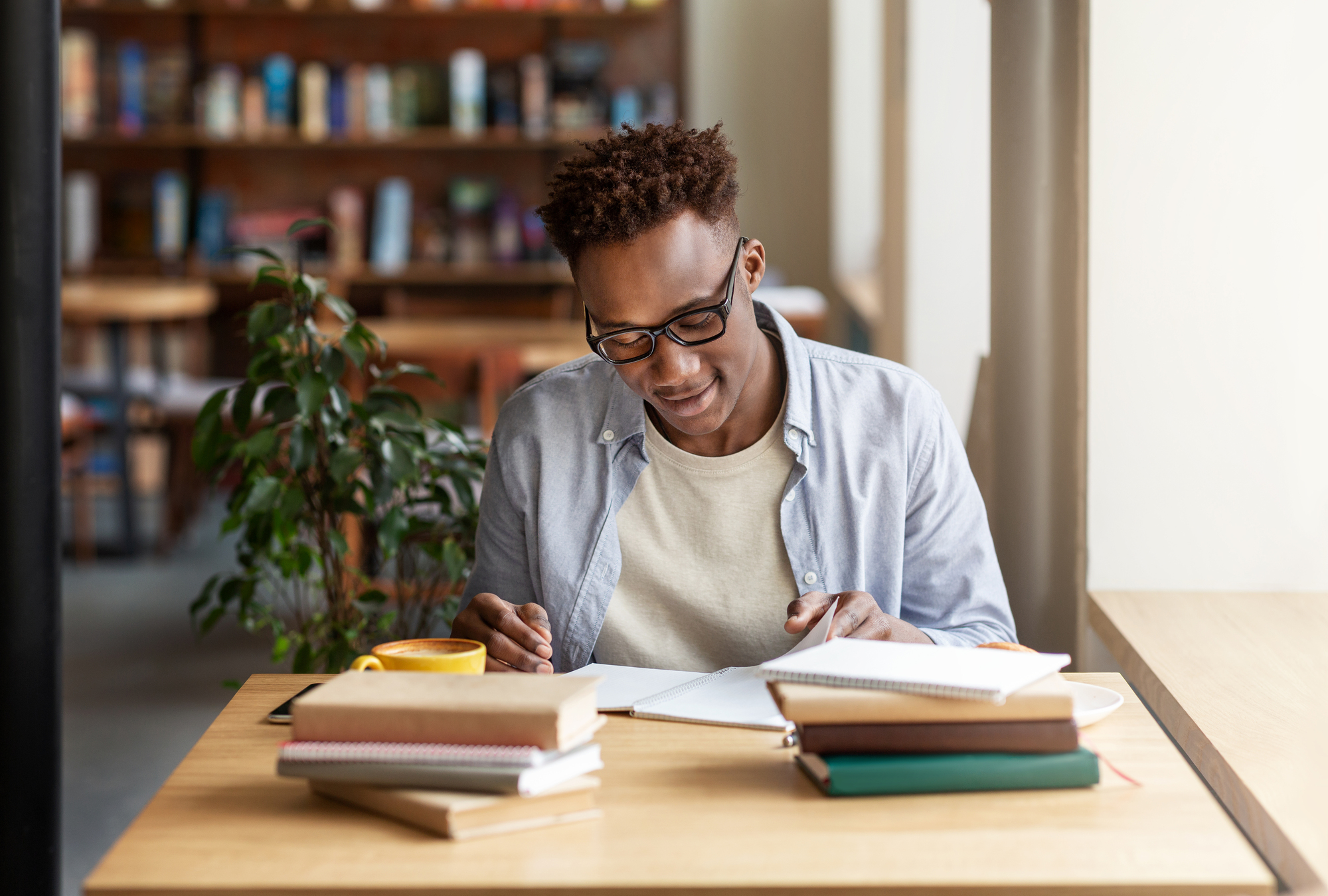 A person wearing glasses studies at a wooden desk with books and a yellow coffee cup. They are focused on writing in a notebook. A potted plant and bookshelves are in the background. The scene is bright and well-lit.