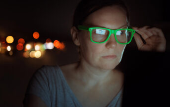 A person with short hair and neon green glasses looks at a screen in a dark setting. The screen light reflects off their glasses. Out-of-focus streetlights in the background create a bokeh effect.